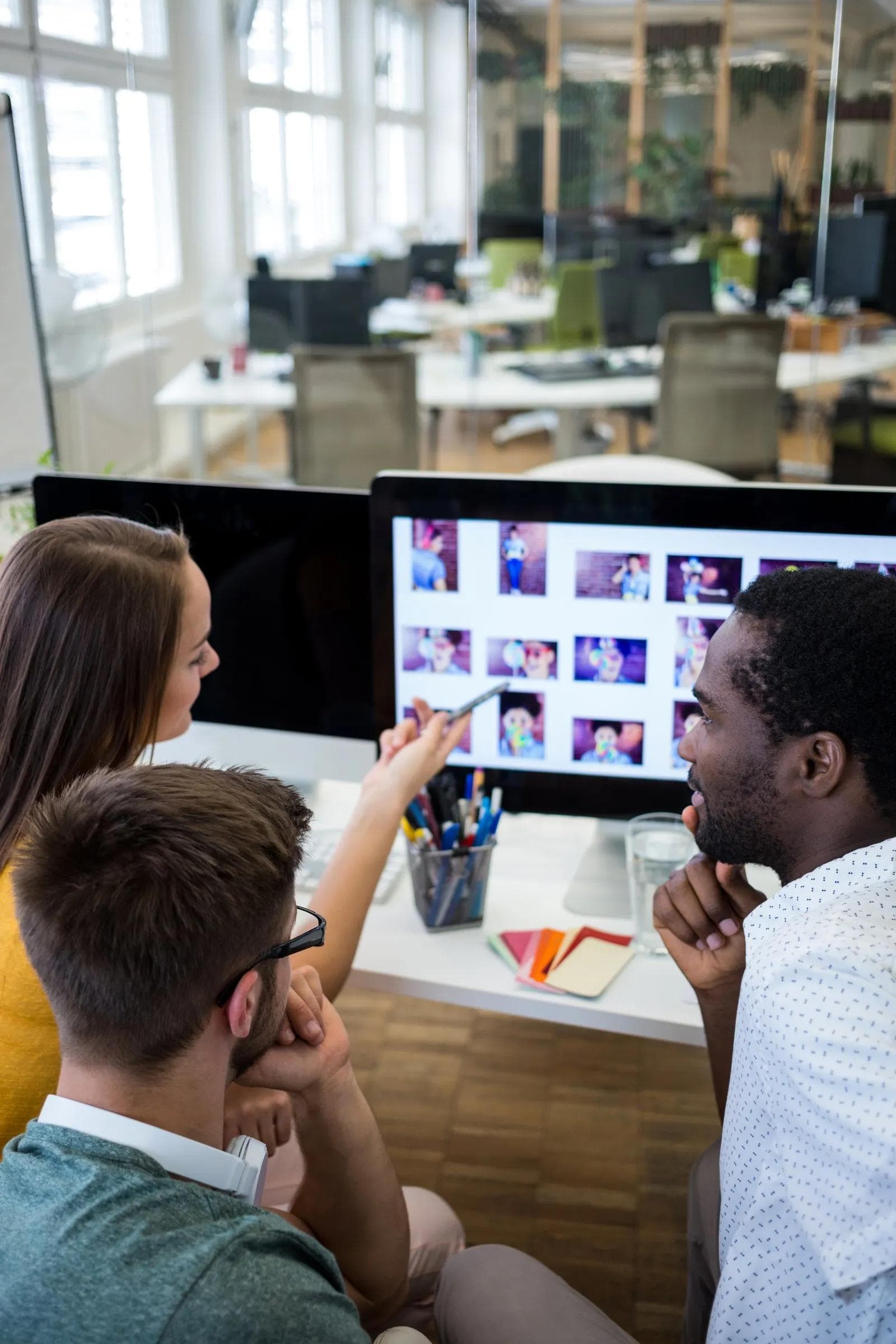 Three colleagues in an open-plan office reviewing a grid of images on a desktop monitor, one of them pointing at the screen with a pen.
